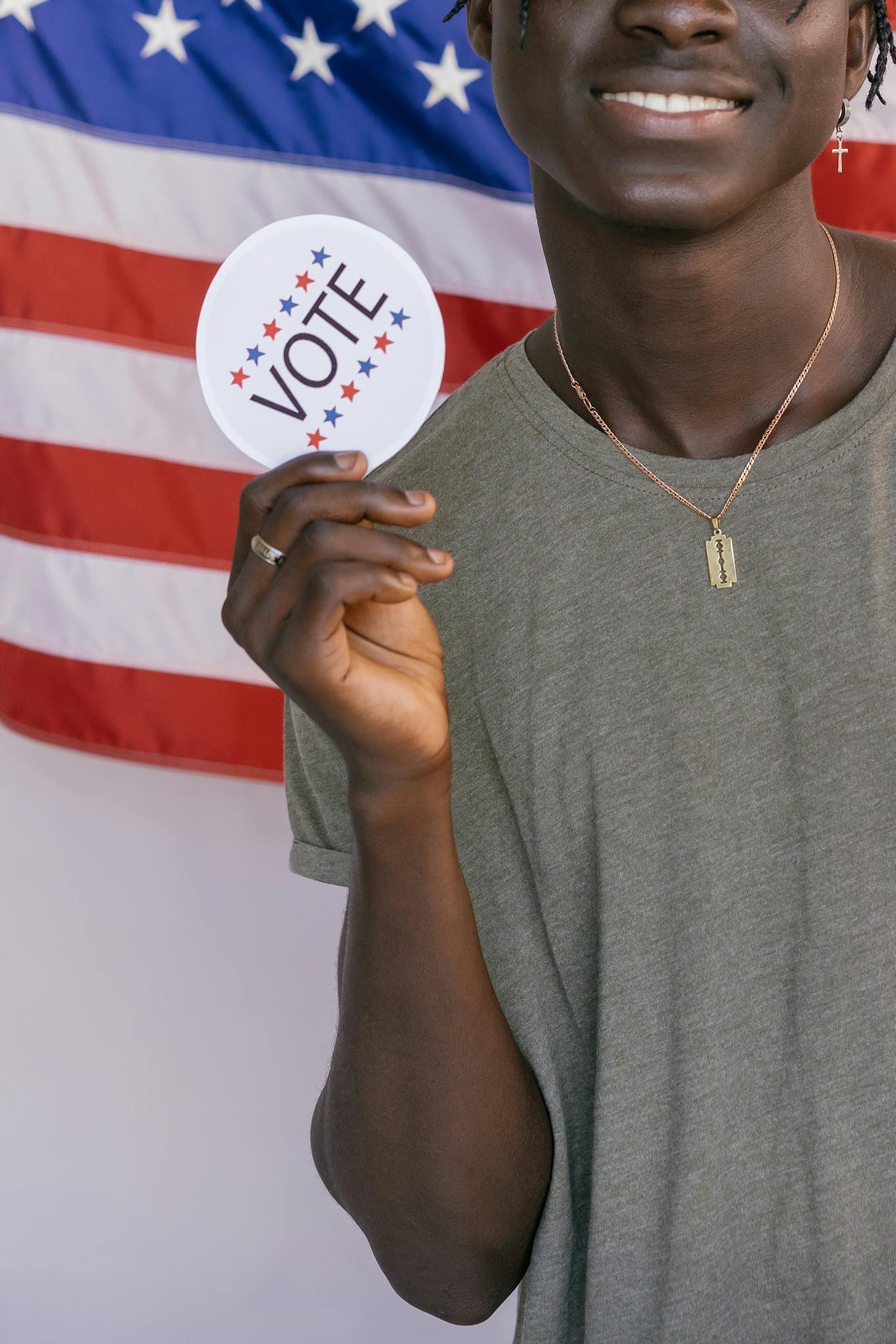 Half-visible person's hand holding a 'Vote' sticker in front of an American flag, symbolizing democratic participation.